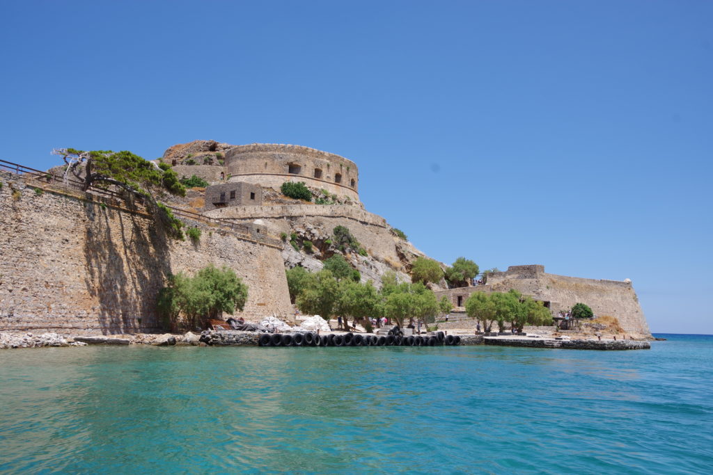 Im Vordergund türkisblaues Meer, im Hintergrund Bootsanlegestelle unter einer Festung bei blauem Himmel