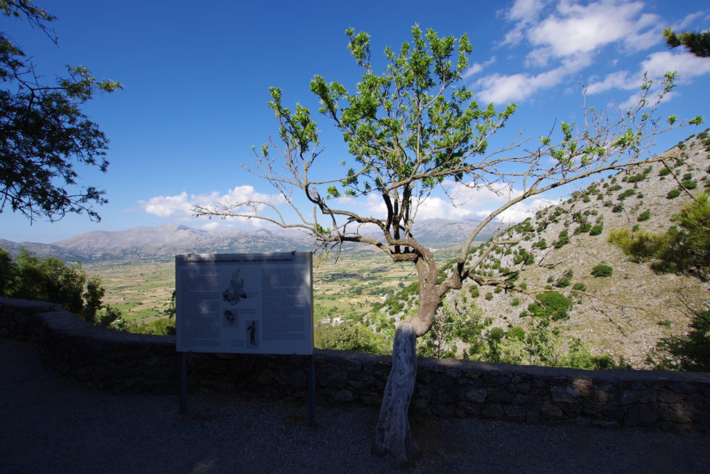 Im Vordergrund einzelner Baum neben Infotafel. Im Hintergrund Hochebene, umrahmt con Bergen unter blauem Himmel