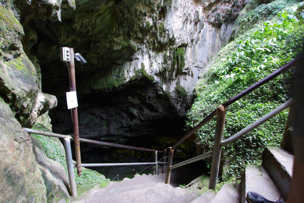 Blick nach unten in den Eingang einer Sackloch-artigen Höhle. Treppenstufen mit Geländer führen hinab