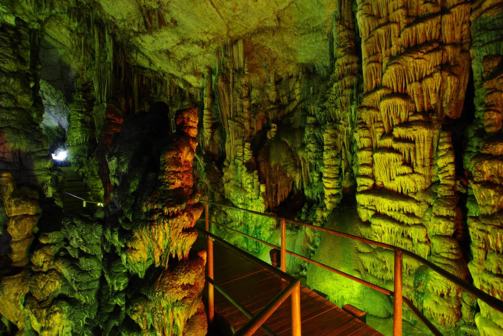 Höhlenraum mit schönen Tropfsteinen, rötlich und grünlich erscheinend. Steg mit Geländer verläuft zwischen Stalagmiten