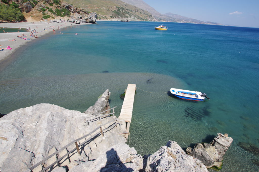 Blick von oben auf Badestrand und Bootsanlegestelle mit klarem Meerwasser.