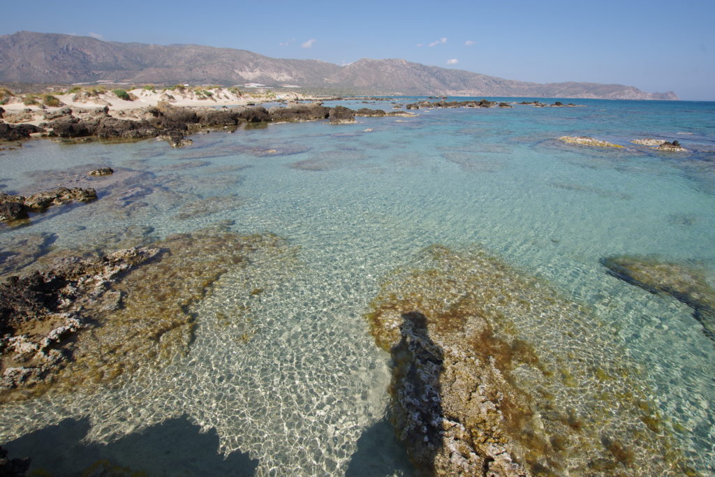 Blick auf seichtes, hellblaues Meerwasser, dahinter Sanddünen und Berge