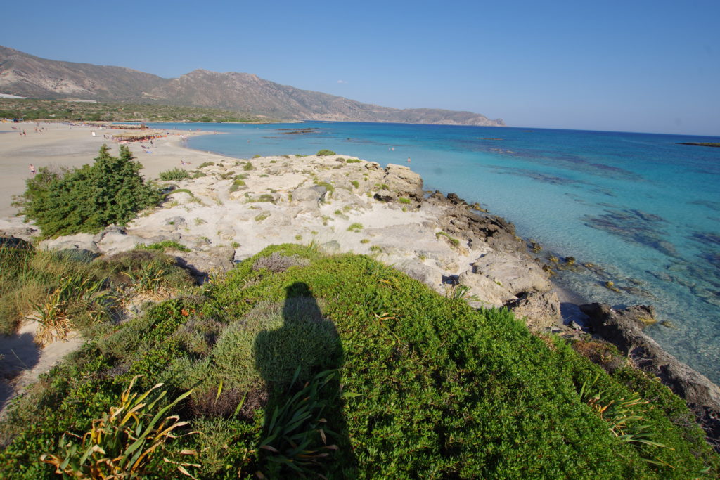 Blick auf Sandbank mit Sonnenschirme, Liegestühle und Touristen, daneben türkis-blaues Meer. Im Vordergrund Dünen mit grünen Sträuchern.