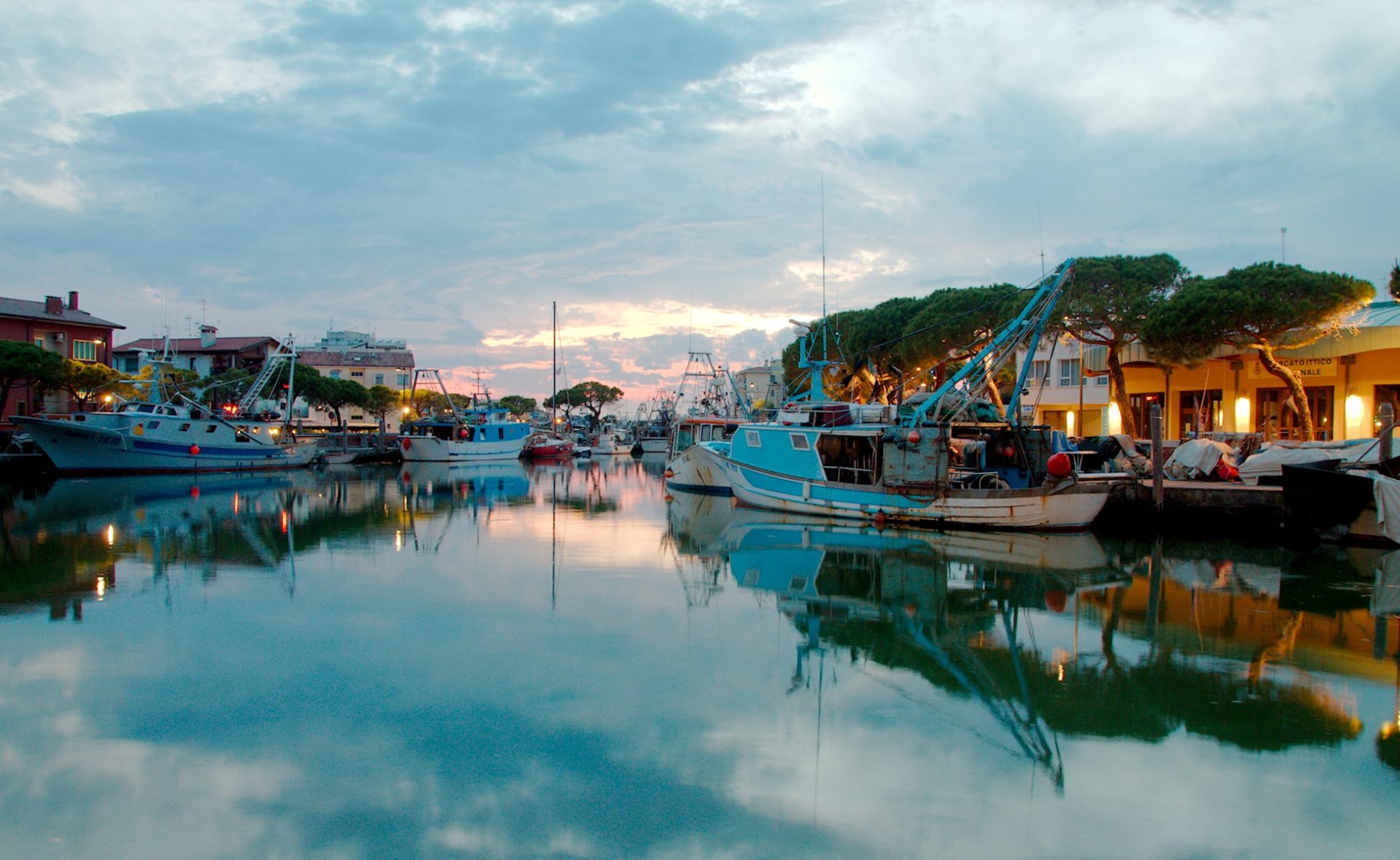 Wassertemperatur Caorle: Hafen in der Dämmerung, szenisch beleuchtete Promenade, romantischer Flair. Wolken und Boote spiegeln sich im stillen Meerwasser.